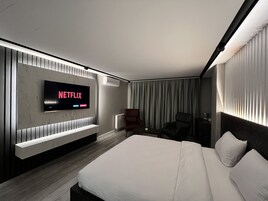 Habitación de hotel moderna con una cama grande, un televisor de pantalla plana montado en la pared, un área de descanso con una silla y una ventana con cortinas.