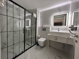 A modern bathroom with a glass shower enclosure, a marble-tiled floor, a white sink with a mirror above it, and a toilet.