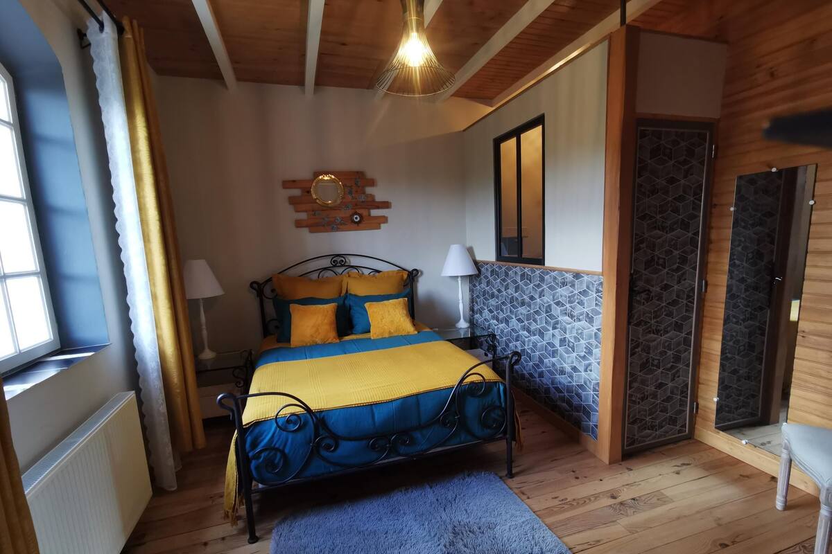 Une chambre à coucher avec un lit, des lampes de chevet, un plancher en bois, un tapis bleu, une fenêtre avec des rideaux et un miroir fixé au mur.