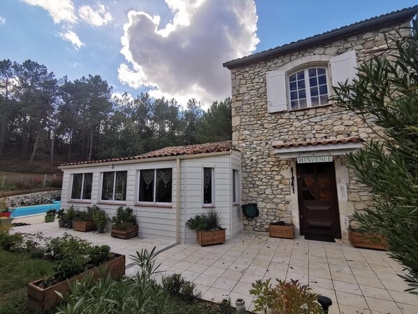A stone building with a tiled roof and a wooden deck. There is a sign that reads "BIENVENUE" on the front door. - Domaine des 3 Cyprès Chez Nath & Pat (Fumel)
