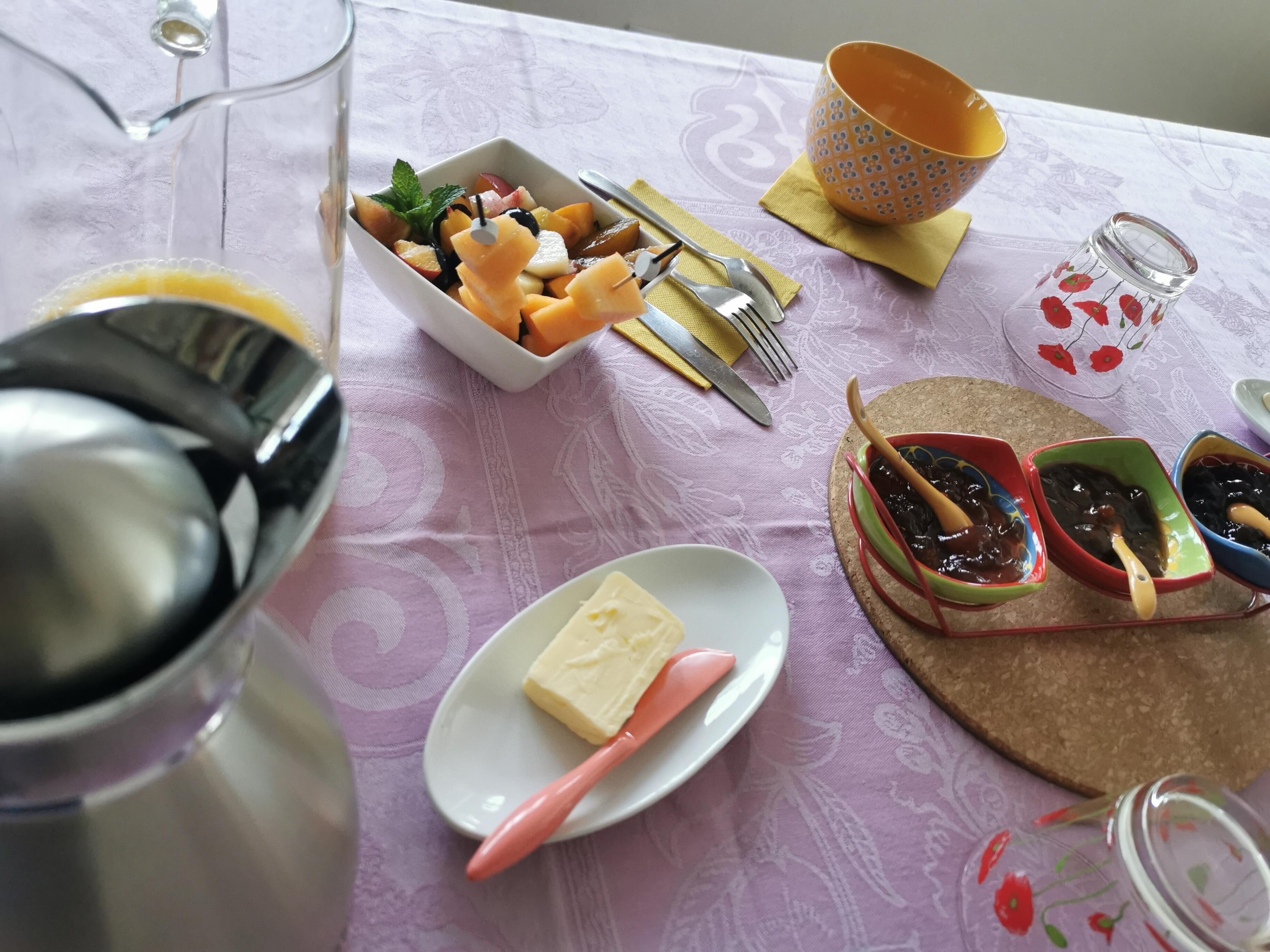 Pöydällä on monenlaista syötävää, kuten kulho täynnä hedelmiä, lautanen, jossa on voita, ja lasinen kannu.