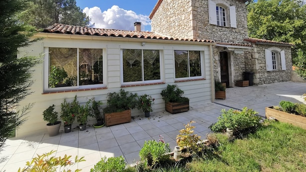 Una casa de piedra de dos pisos, con techo de tejas, ventanas amplias y un jardín con plantas en macetas.