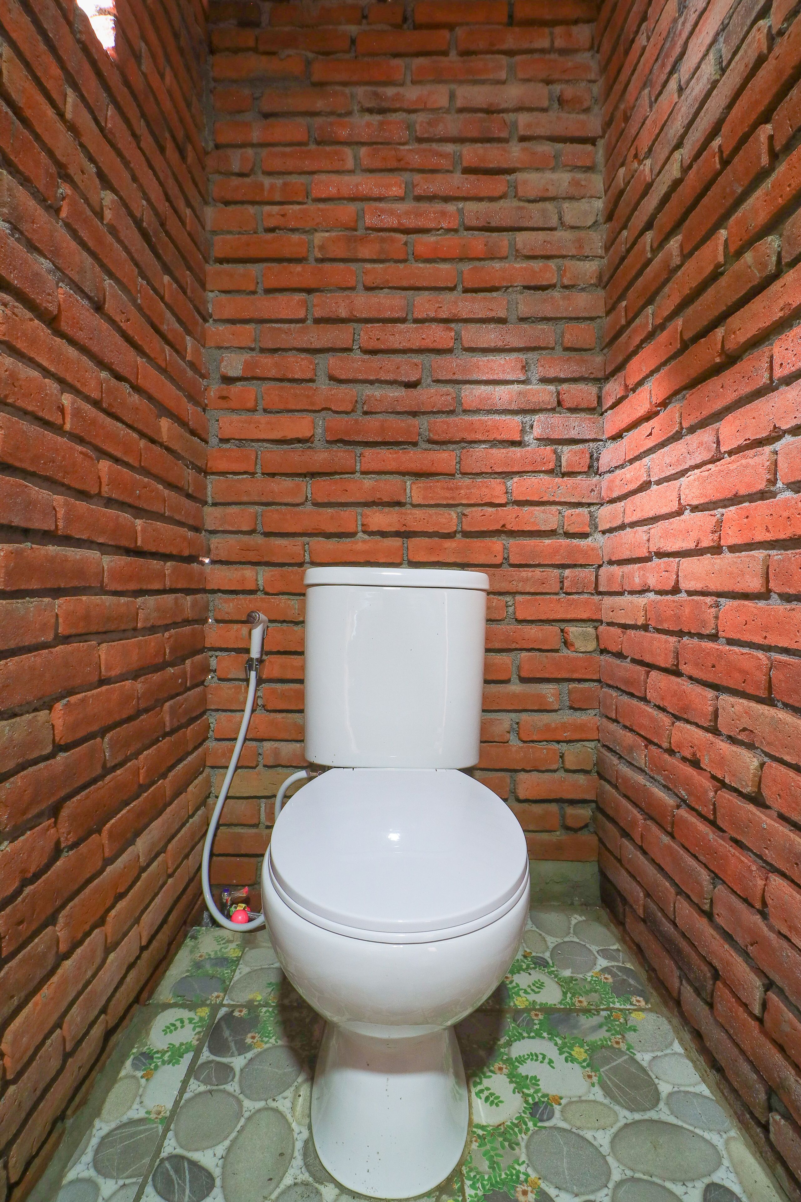Et lille, lukket badeværelse med et hvidt toilet, en synlig rørledning og et mønstret gulv.