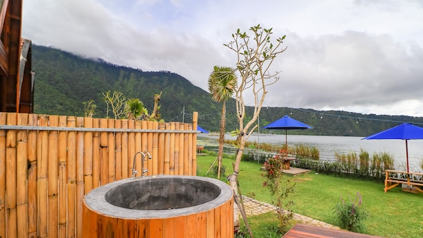 Un jacuzzi en un recinto de madera, un área con césped, un lago y montañas al fondo, y una sombrilla azul.