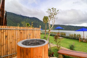Un jacuzzi en un recinto de madera, un área con césped, un lago y montañas al fondo, y una sombrilla azul.