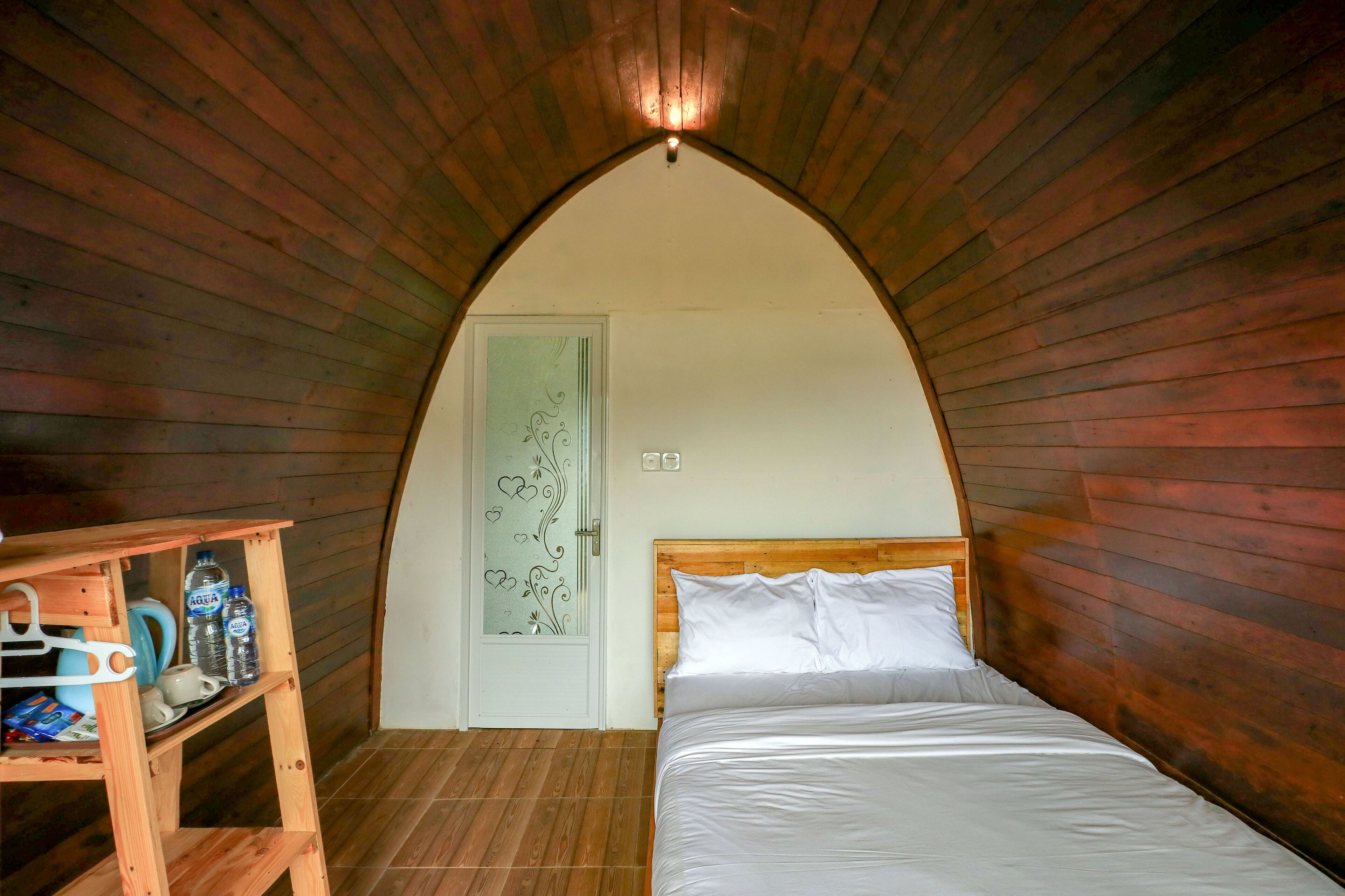 Ein Schlafzimmer mit Holzbalkendecke, einem Bett mit weißer Bettwäsche, einem Nachttisch aus Holz mit einer Tasse und einer Flasche sowie einer Tür mit dekorativer Glasscheibe.