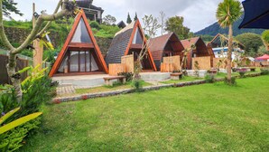 Filas de cabañas de techo triangular con fachada de madera, ubicadas en un exuberante jardín con montañas al fondo.