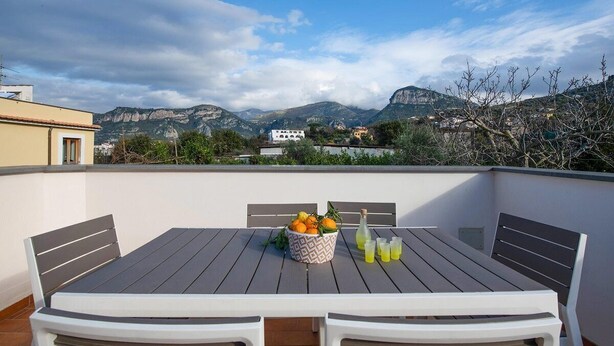 A rooftop terrace with a table set for two, a bowl of fruit, and a bottle of drink.