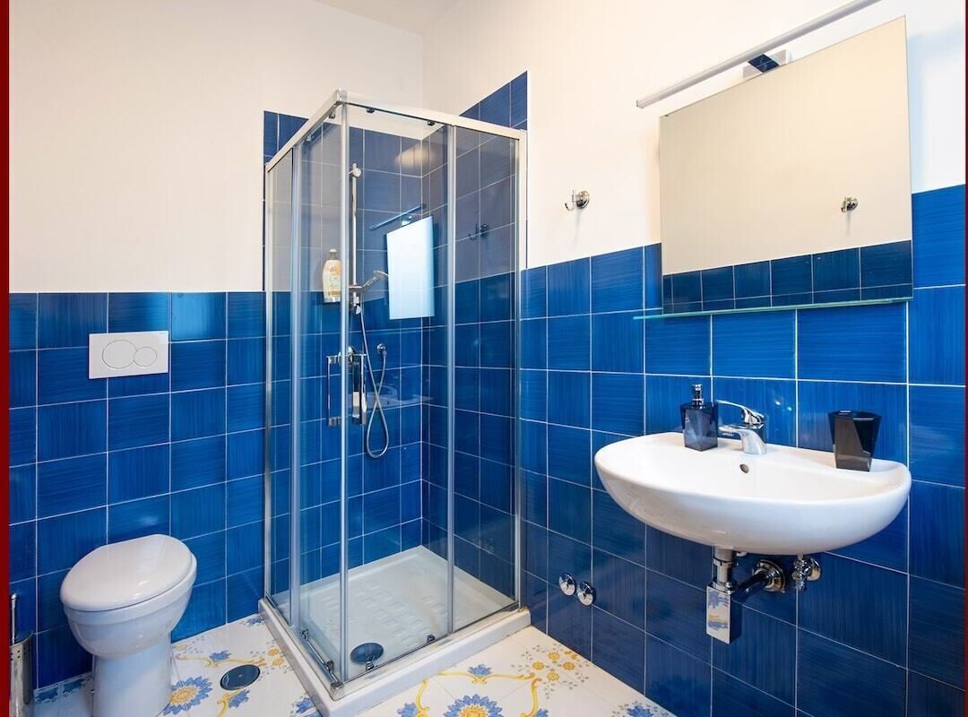Ein Badezimmer mit blauen Fliesen, einer Duschkabine aus Glas, einem weißen Waschbecken und einer Toilette.