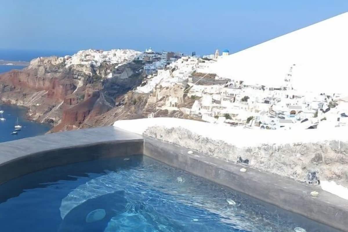 A swimming pool with clear blue water, surrounded by a stone deck. In the background, there is a view of a coastal area with white buildings and rocky cliffs.