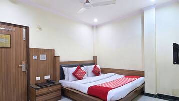 A hotel room with a wooden wardrobe, a bedside table with a phone and a book, a bed with white bedding and red pillows, and a ceiling fan.