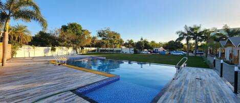A swimming pool with a wooden deck, surrounded by palm trees and a clear sky.