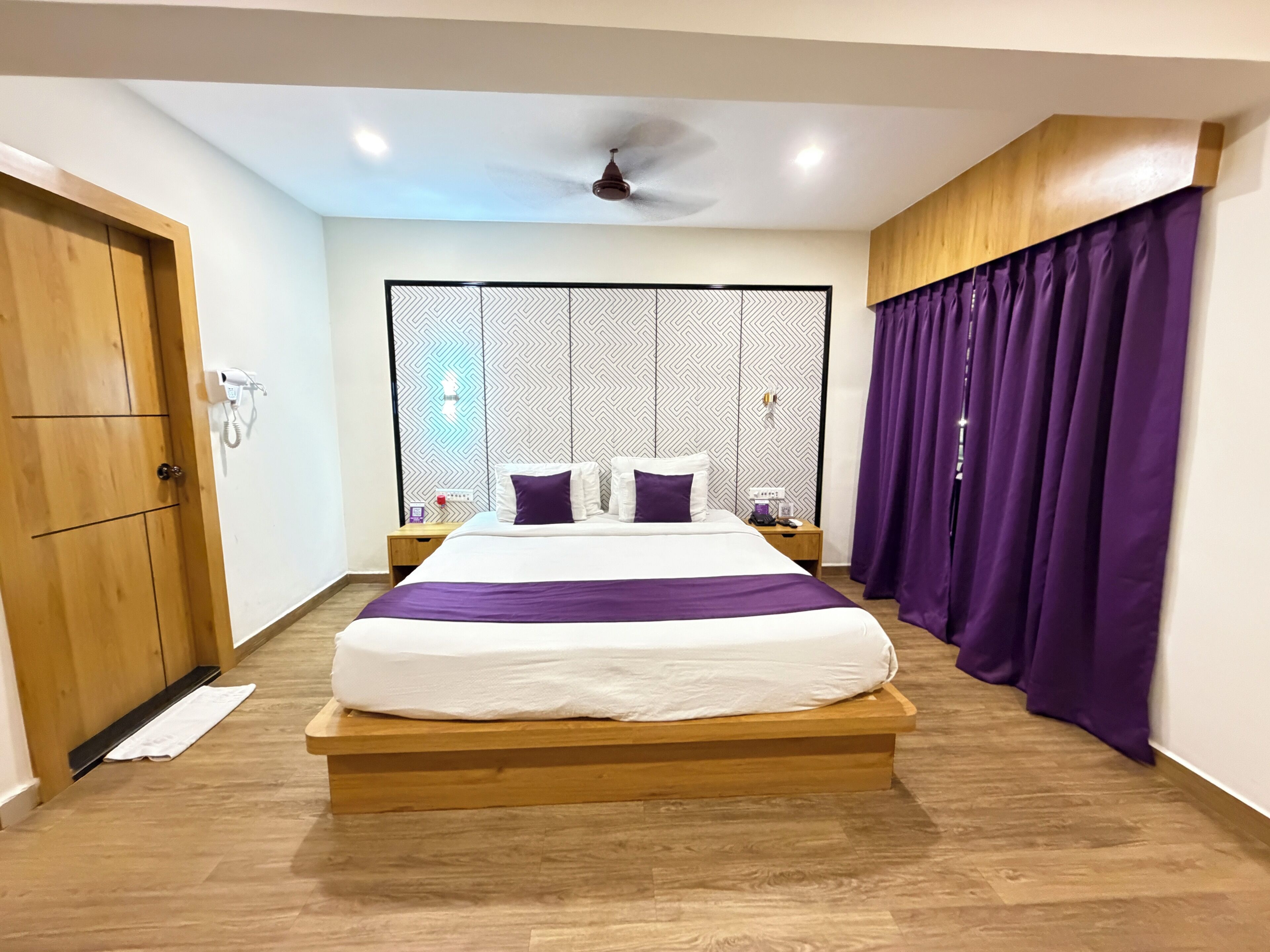 Habitación de hotel con cama, cabecera de madera, cortinas moradas, ventilador de techo y un separador de ambientes con diseño.