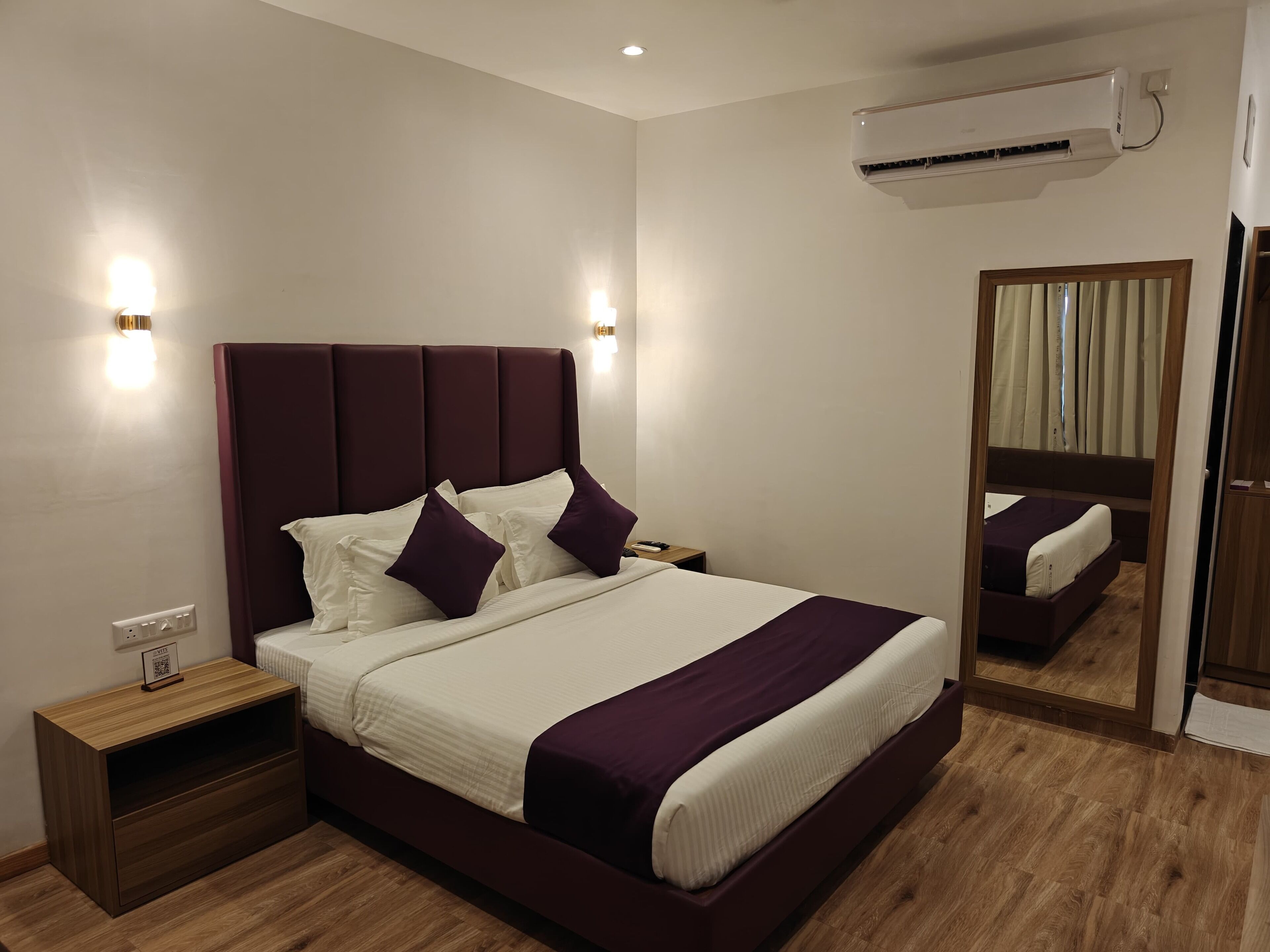 A modern bedroom with a large bed, bedside tables, an air conditioning unit, and a mirror.