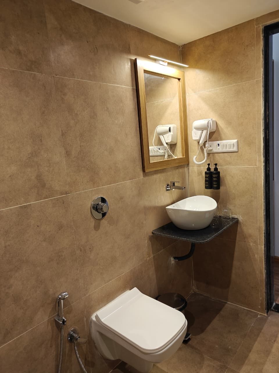 Un baño con inodoro blanco, lavamanos con recipiente, secador de pelo de pared y ducha con cabezal de ducha manual.