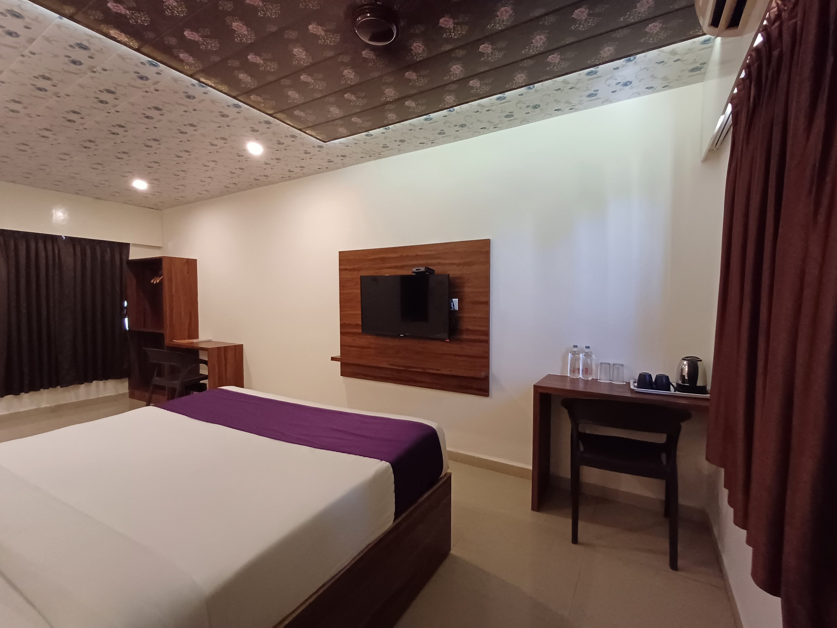 A hotel room with a bed, a desk, a TV mounted on the wall, and a ceiling with a floral pattern.