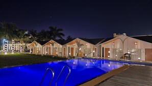 A nighttime view of a resort with a swimming pool, wooden deck, and multiple villas with arched doorways and large windows. - VITS Daman Devka Beach (Daman)