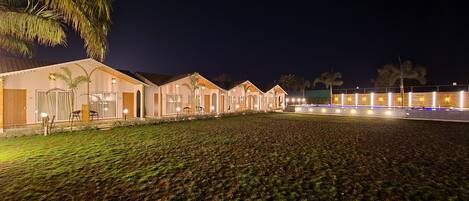 A row of well-lit, single-story buildings with large windows, a swimming pool, and a grassy area in the foreground.