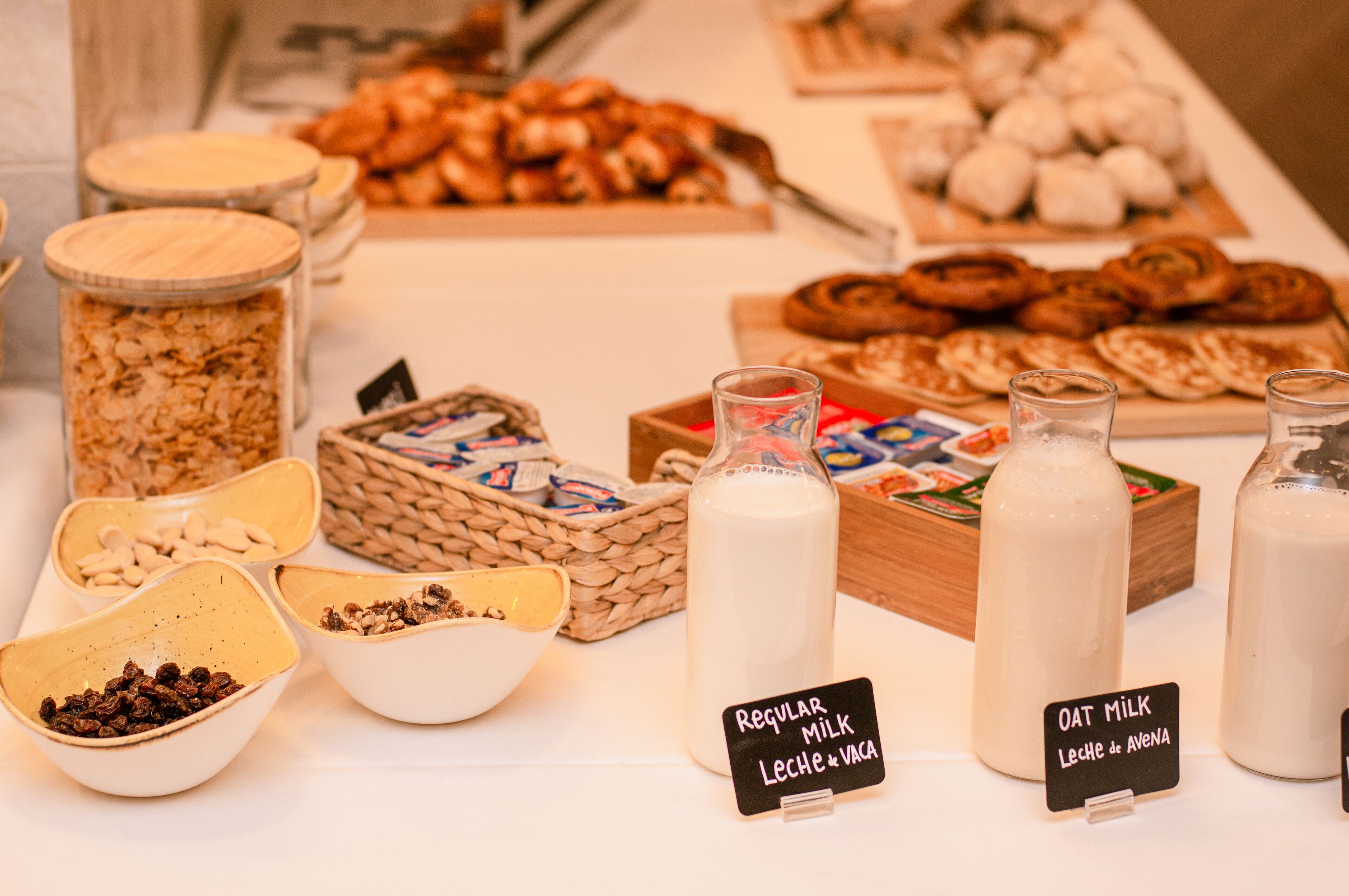 Una mesa de buffet con diversos alimentos y botellas de leche, algunas etiquetadas como 'Regular Milk' y 'Oat Milk'.