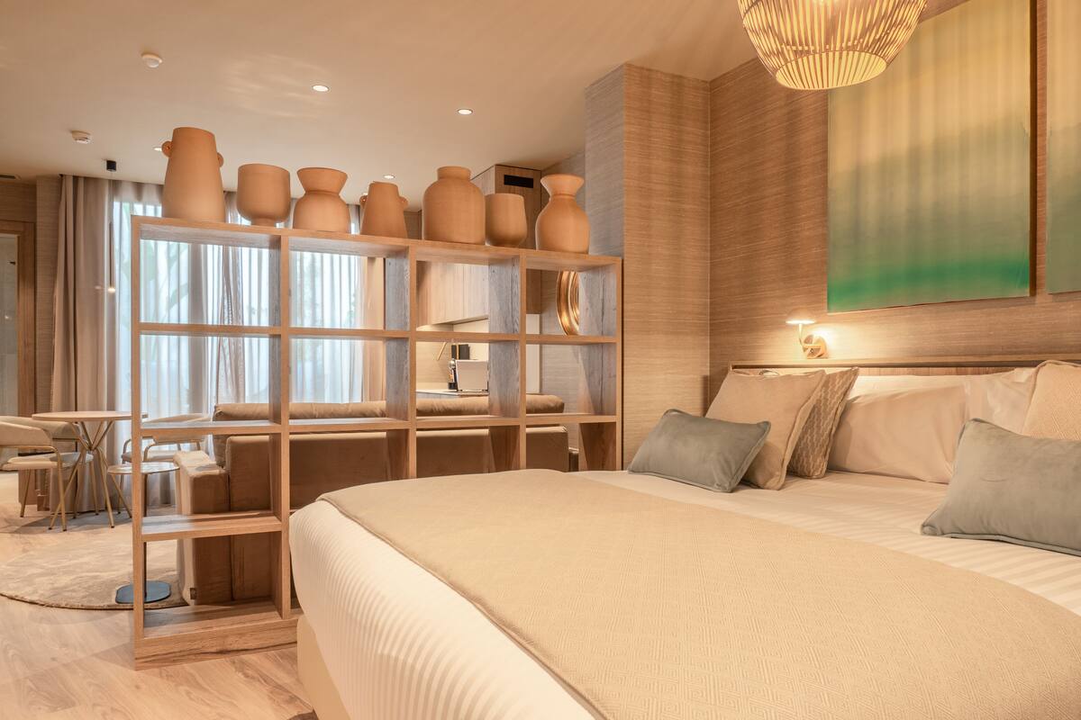 A modern hotel room with a large bed, a wooden shelf with decorative vases, a dining area, and a large window with curtains.