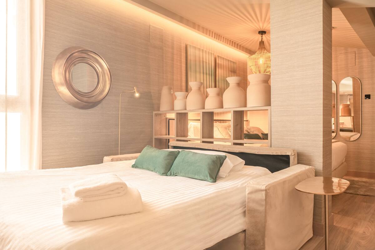 A modern hotel room with a neatly made bed, a bedside table, a mirror, and a wall-mounted shelf with decorative items.
