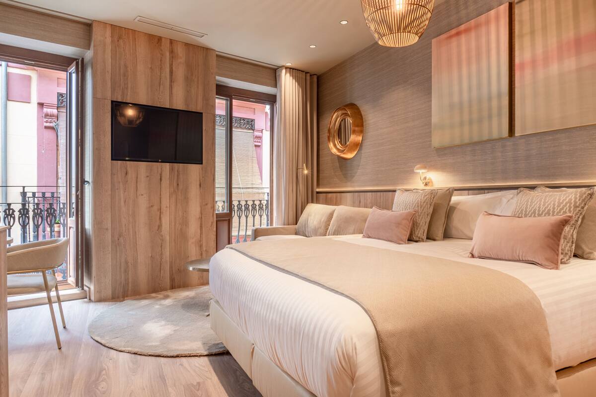 A modern bedroom with a large bed, a TV mounted on the wall, and a balcony with a view of the city.
