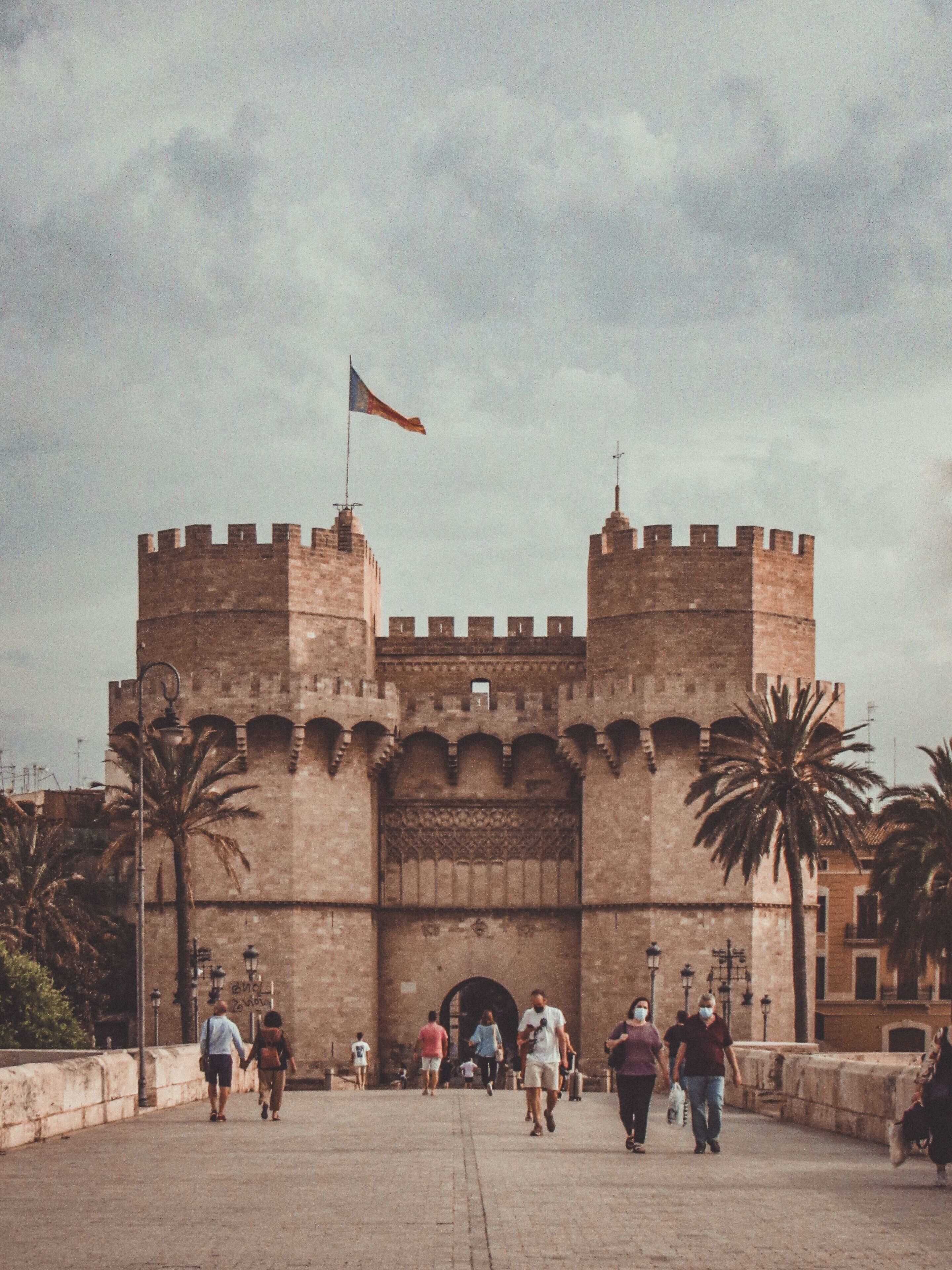 A historic stone gate with two towers, palm trees, and people walking in the foreground.
