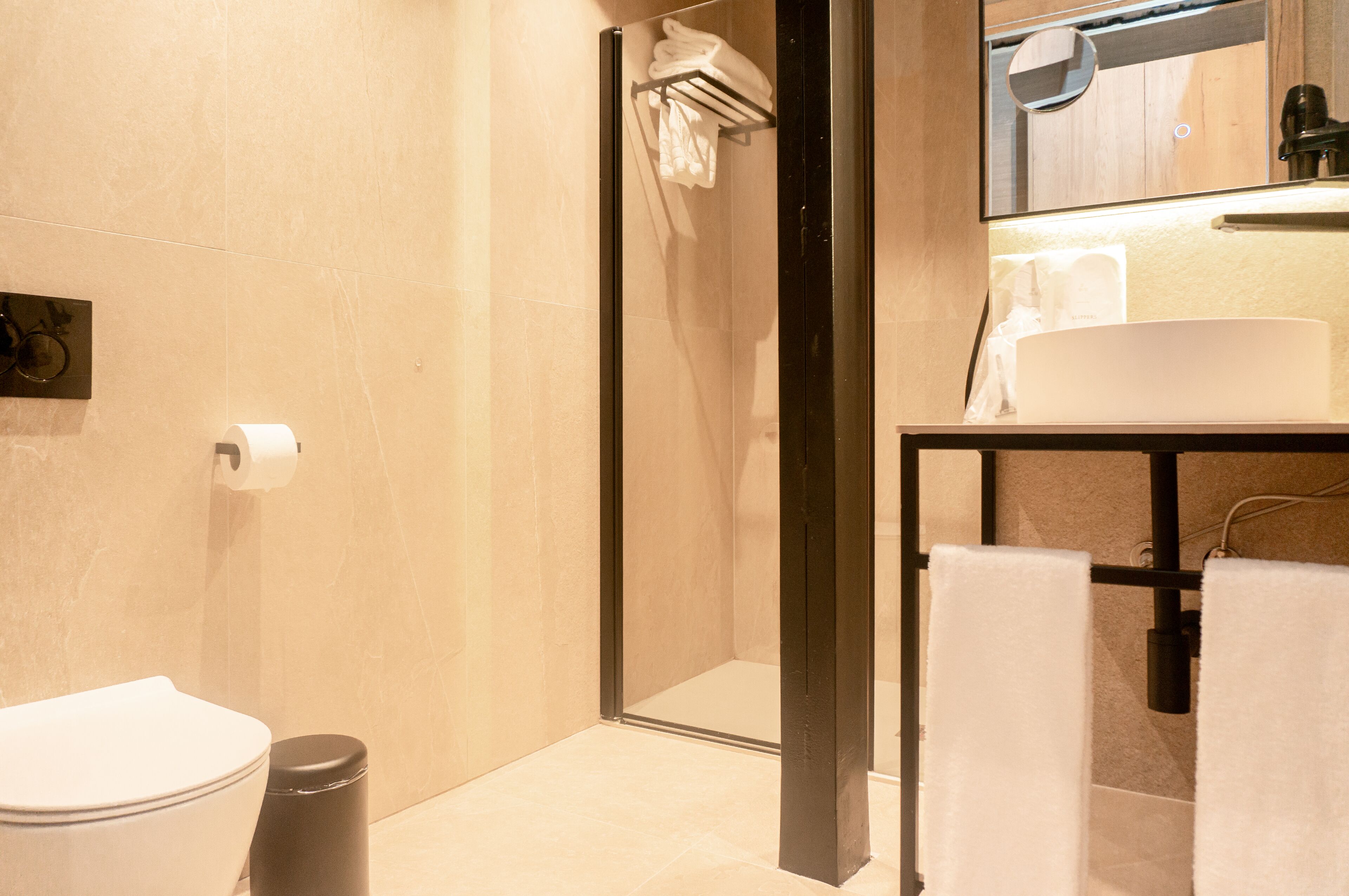 A modern bathroom with a toilet, a trash bin, a shower area, and a sink with a towel rack.