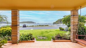 A covered outdoor area with stone pillars, a view of a road and water body, and a grassy lawn.