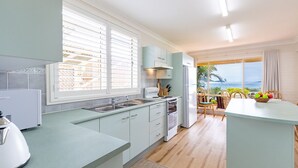 A modern kitchen with light blue cabinets, a large window with blinds, a dining area with a view of the sea, and a small island with fruit. - Anchored at Fingal Bay, 80 Marine Drive (Fingal Bay)
