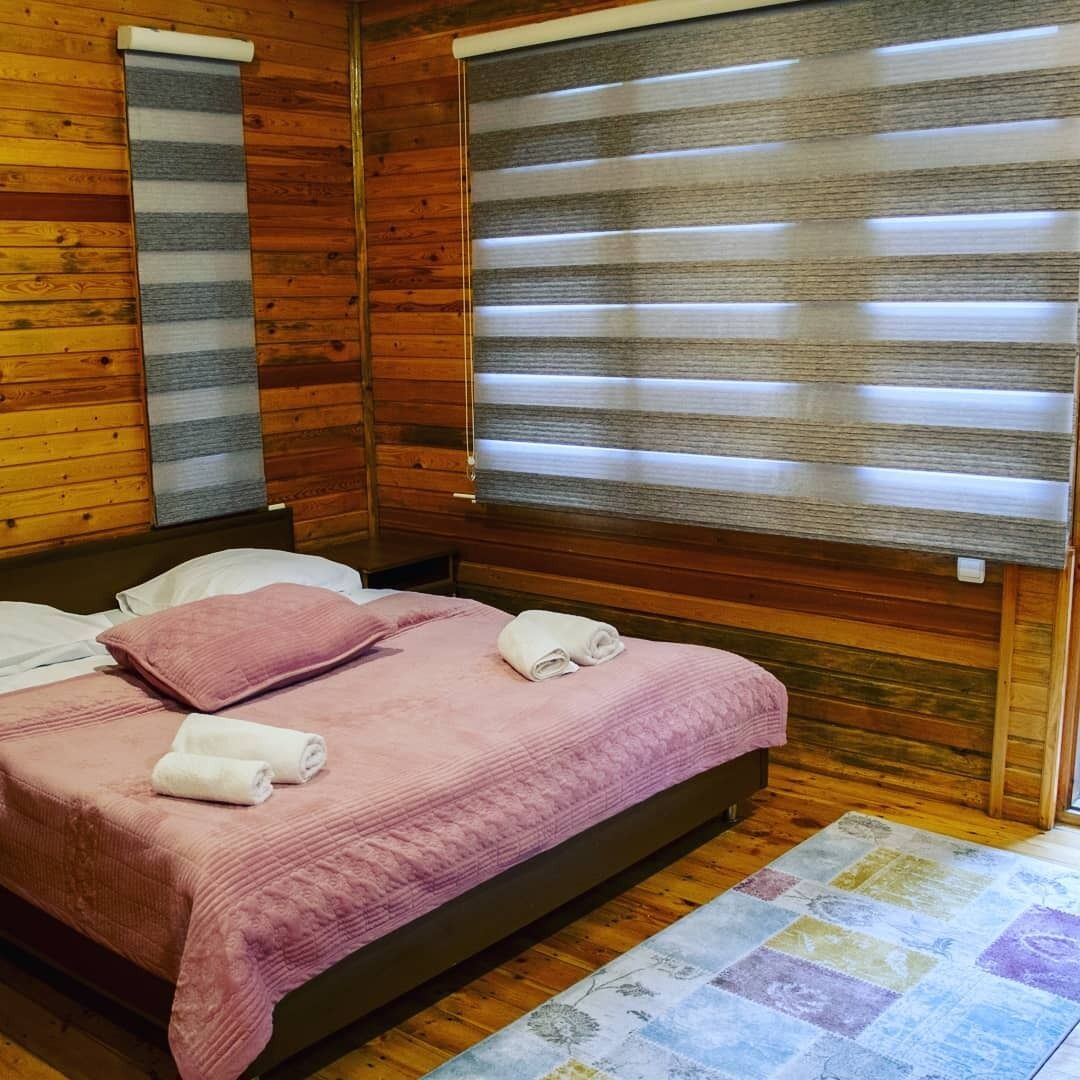 A wooden cabin room with a bed, two windows with blinds, a quilted pink bedspread, and a colorful rug.