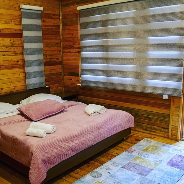 A wooden cabin room with a bed, two windows with blinds, a quilted pink bedspread, and a colorful rug.