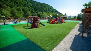 Een speeltuin met diverse speeltoestellen, omringd door groen gras en een beboste heuvel.