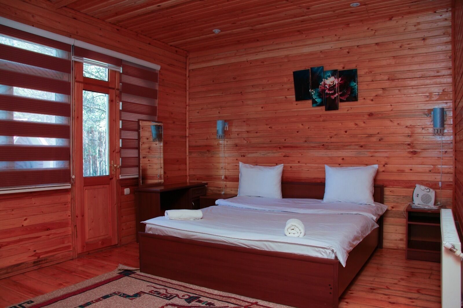 A wooden cabin bedroom with a bed, bedside tables, a mirror, and a window with blinds.