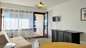 A room with a bed, a round table with a yellow tablecloth, a white chair, a black cabinet, and a TV mounted on the wall.