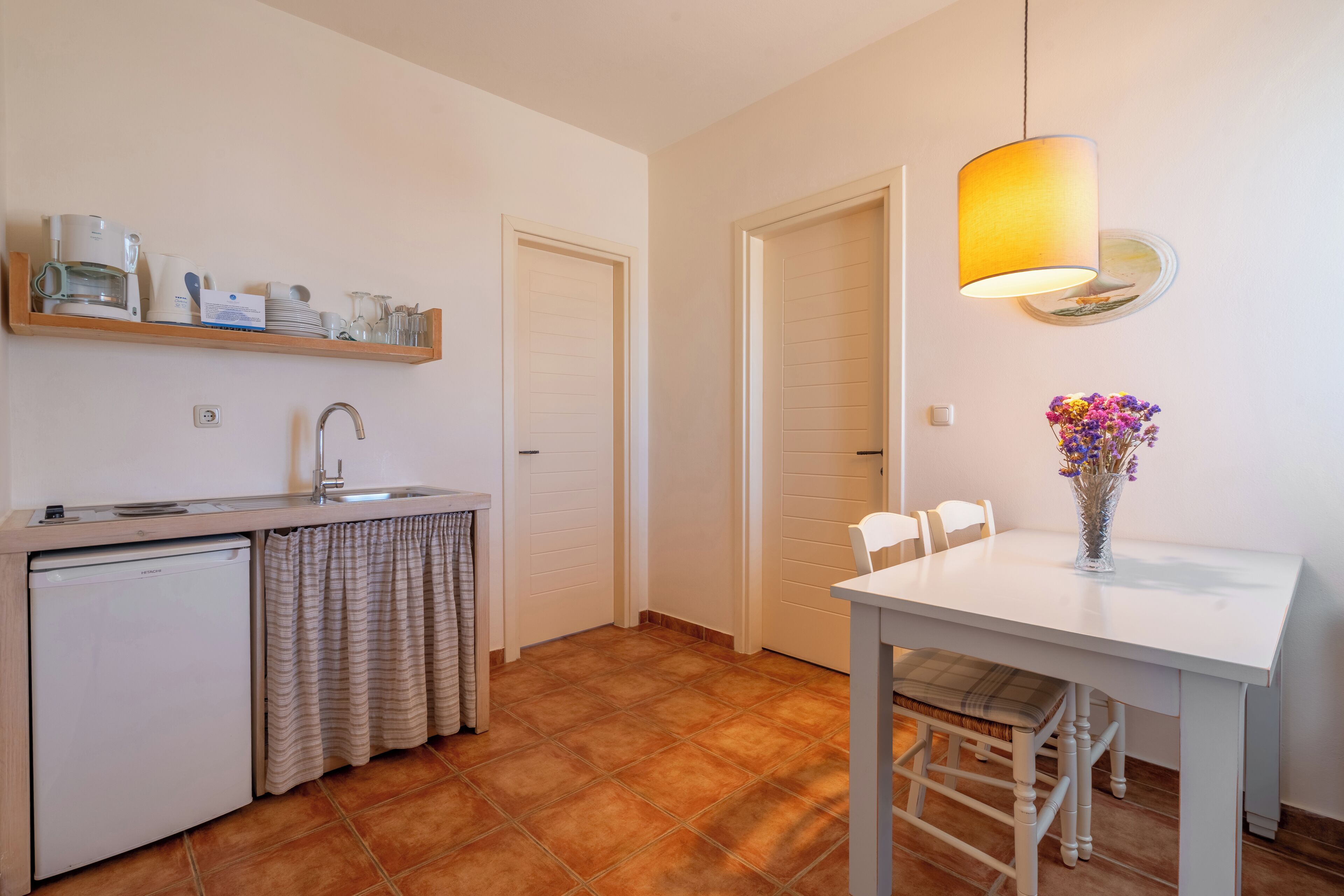 A compact kitchen with a sink, a small refrigerator, a wooden shelf with kitchenware, a dining table with chairs, and a hanging lamp.