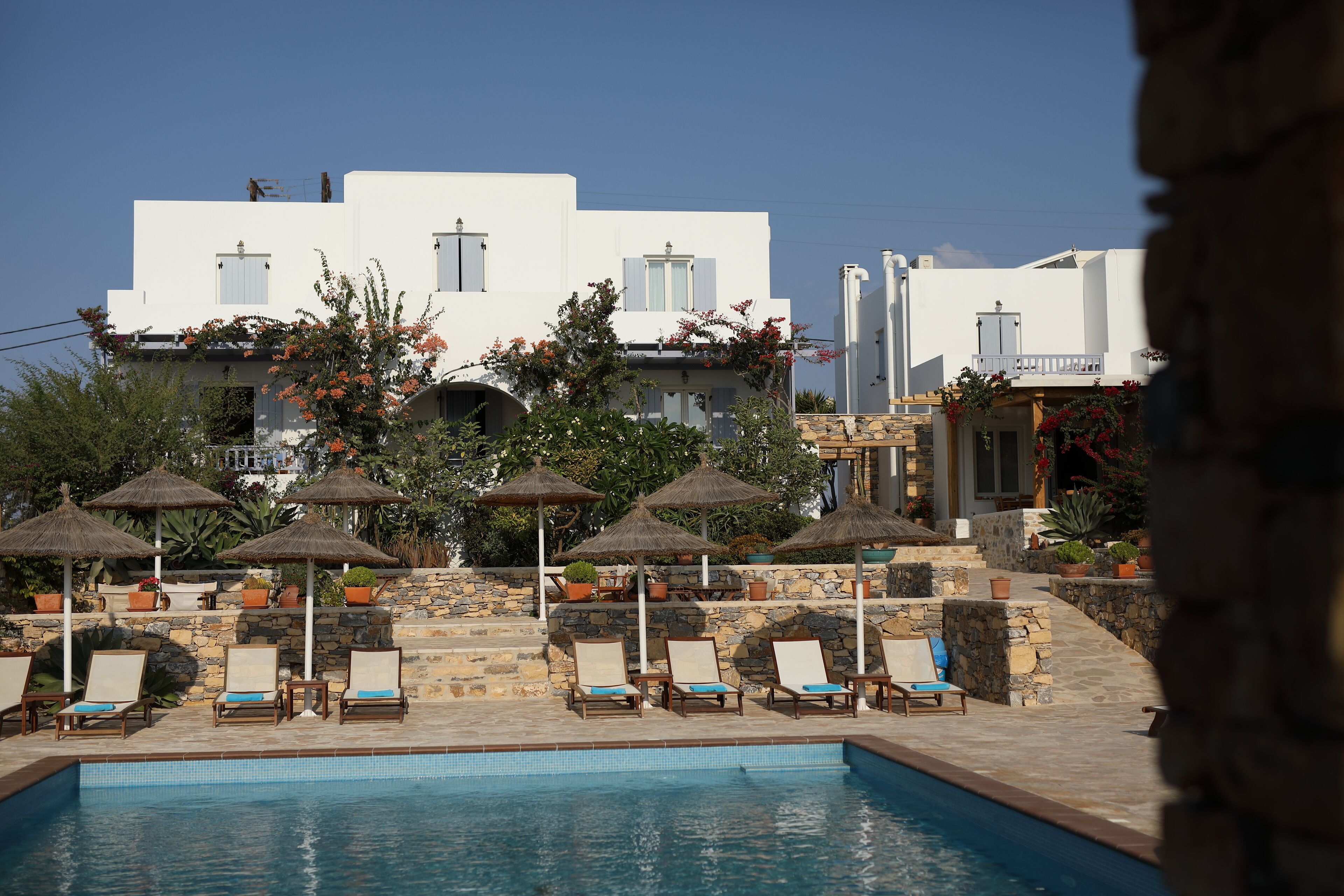 A poolside area with lounge chairs and umbrellas, a stone wall, and a white building with windows and balconies.