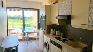 Une cuisine compacte équipée d’un four encastré, d’un lave-vaisselle et d’un évier. On y trouve également un coin repas avec une table et des chaises, et une vue sur un paysage verdoyant grâce à une porte vitrée.