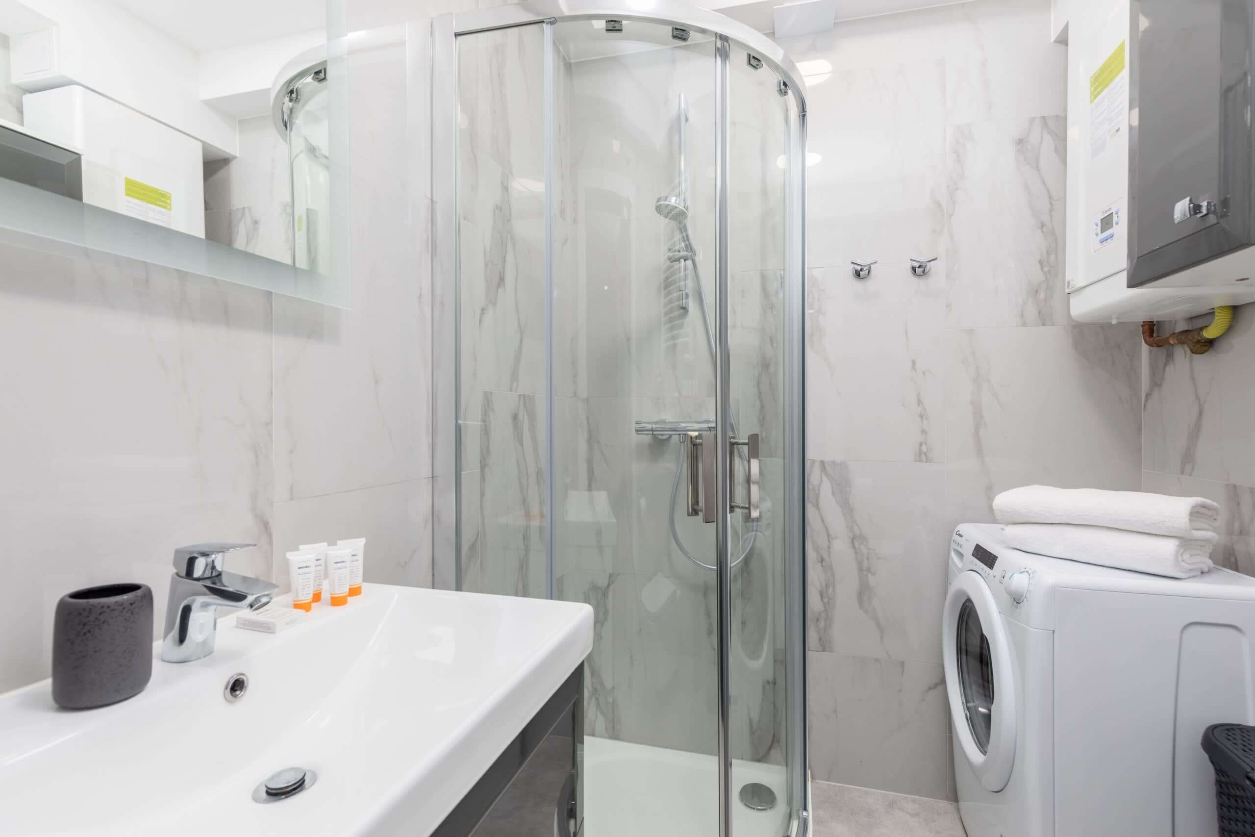 A modern bathroom with a glass shower enclosure, a sink with toiletries, and a washing machine.