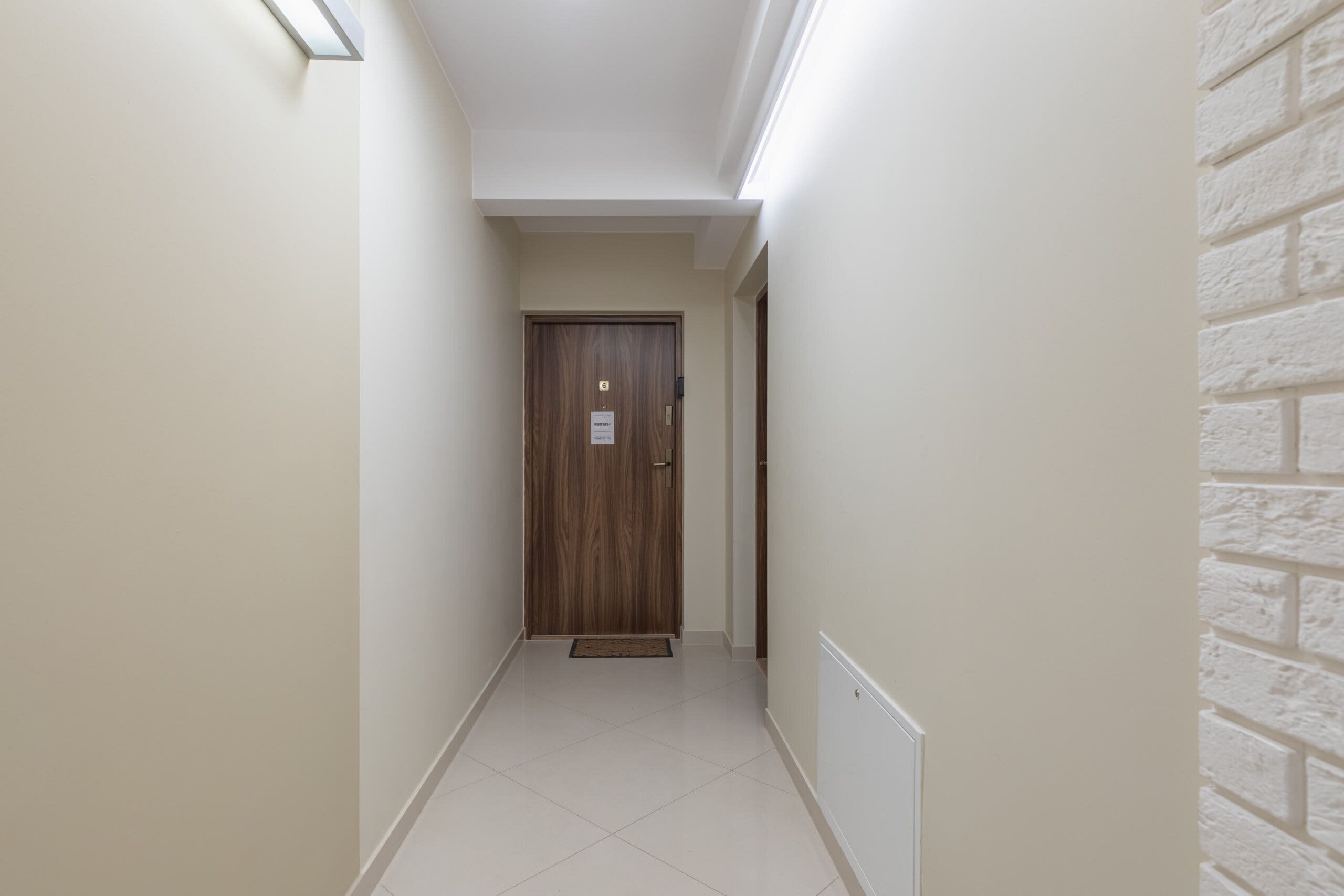 Un pasillo estrecho con una puerta de madera al final, paredes blancas y una pared de ladrillo blanco decorativa.
