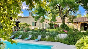 A swimming pool with lounge chairs, surrounded by lush greenery and a stone building with a tiled roof.
