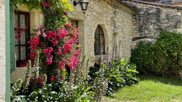 A stone building with a green door and windows, surrounded by lush greenery and flowering plants.