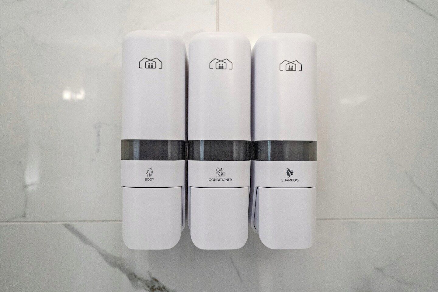 Trois distributeurs blancs, placés sur une surface carrelée, et portant les inscriptions « CORPS », « APRÈS-SHAMPOING » et « SHAMPOING ».