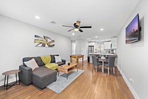 A modern living room with a sofa, coffee table, and a foosball table. There is a dining area with chairs and a table. The room has hardwood floors and white walls.