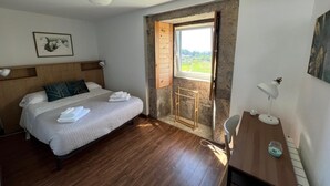 Ein Schlafzimmer mit einem hölzernen Bett, einem Nachttisch, einem Schreibtisch mit Lampe und einem Fenster mit Blick ins Grüne.