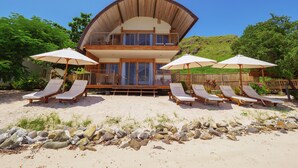 Una villa frente al mar de dos pisos con techo curvo, varias sillas de descanso y sombrillas.