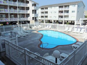 A swimming pool surrounded by white railings and lounge chairs, with multi-story buildings in the background.