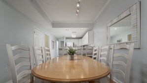A dining area with a round wooden table surrounded by white chairs, a potted plant on the table, and a large mirror on the wall.