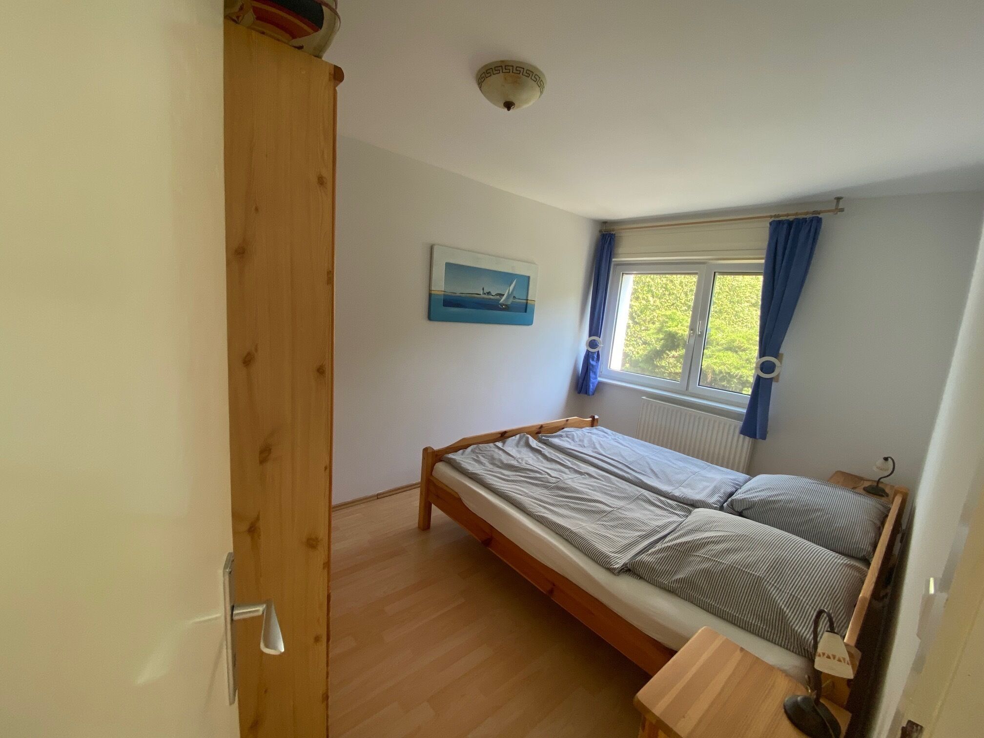 A bedroom with a single bed, a wooden wardrobe, a window with curtains, and a ceiling light fixture.