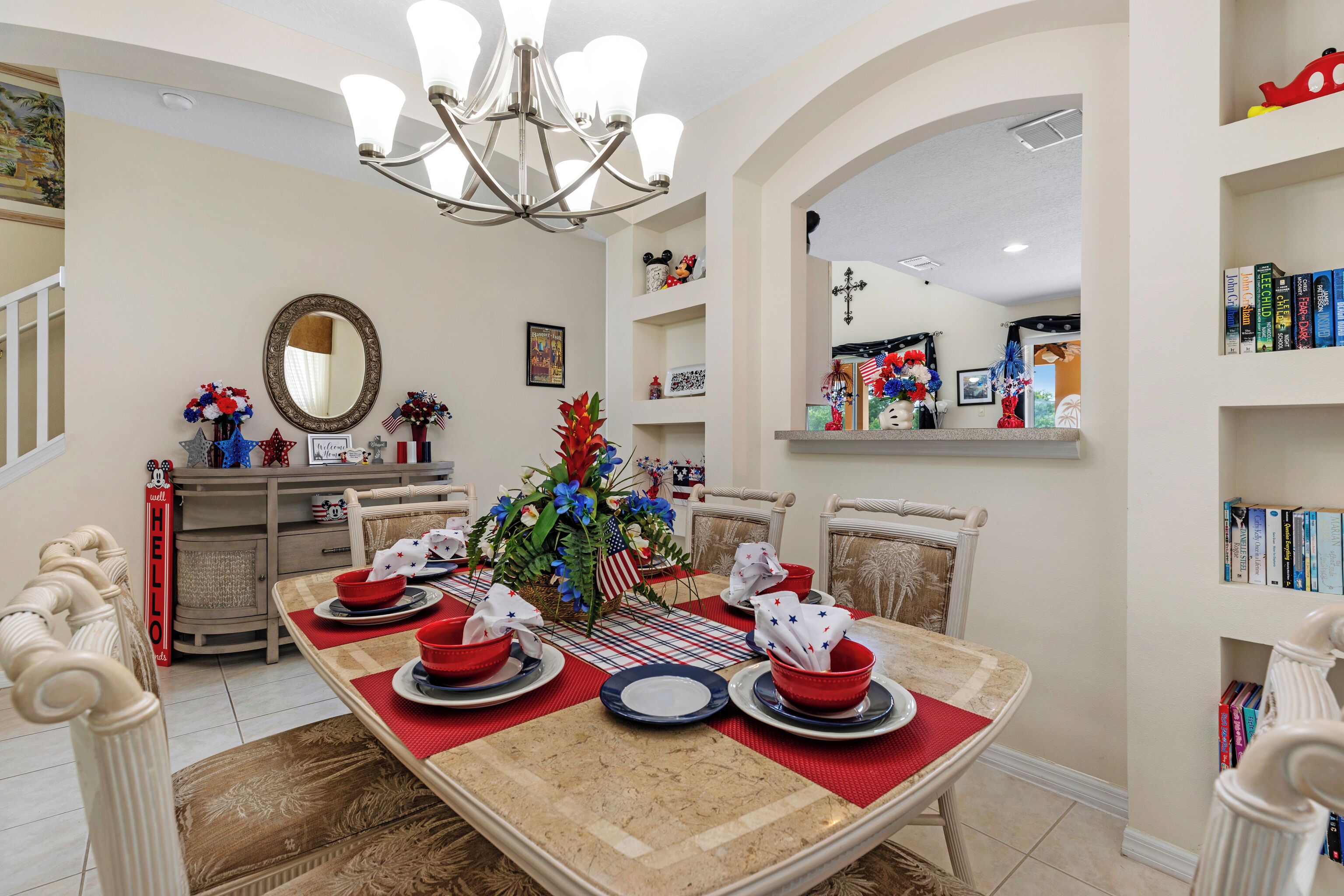 Uma sala de jantar com uma mesa grande posta para quatro pessoas, um lustre e uma estante repleta de livros.